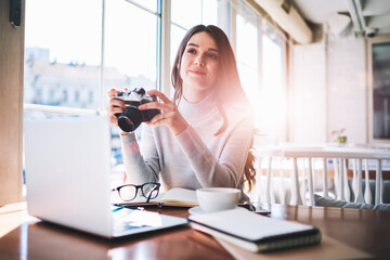 Young happy brunette hipster girl sitting at cozy cafeteria holding vintage camera in hands while waiting friend, caucasian female photographer spending time at coworking space and looking away