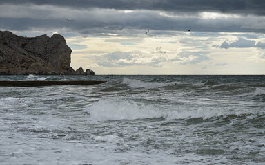Seascape on shore of the Black Sea in eastern Crimea. White wave crests and bubbling water in the foreground. Dark clouds and three seagulls in the sky over Mount Alchak. Light clouds on horizon.