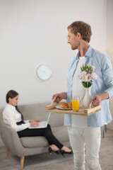 Thoughtful guy bringing his girlfriend food on tray. Handsome middle aged man in glasses holdin tray with ornage juice and croissants for his girlfriend.