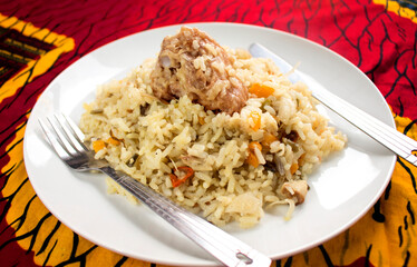 A tasty white plate of Nigerian fried rice containing sliced carrots, green beans and peppers with a piece of turkey meat on top. A fork and knife is placed on the side with a colorful red table cloth