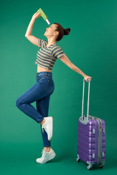Happy young Asian woman holding suitcase, passport and flight ticket while standing isolated over green background.