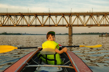 A boy with his father is kayaking on the river near the bridge in the summer.