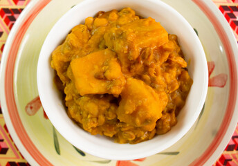 A white bowl of Yam and Beans Pottage or Nigerian Porridge served on a Plate with a colorful red pattern African tablecloth from top view