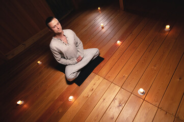 A portrait of an attractive Caucasian man sits in cotton robes in a lotus pose in a dark wooden...