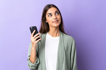 Young caucasian woman using mobile phone isolated on purple background looking up while smiling