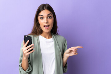 Young caucasian woman using mobile phone isolated on purple background surprised and pointing...