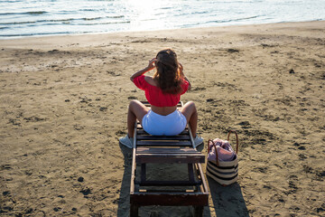 Beautiful girl in a headphones in a shorts and blouse is relaxing on wooden lounge chair on a sandy beach by the sea. Top view. Sunset.