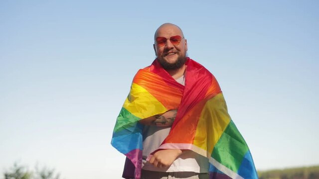 Homosexual bold man wrapped in a rainbow gay flag while parade on background of blue sky. LGBTQI, Pride Event, LBGT Pride Month, Gay Pride Symbol