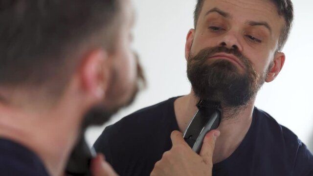 Man shaves his beard using an electric trimmer. Reflection in the mirror