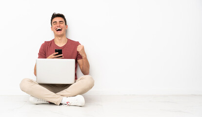 Teenager man sitting on the flor with his laptop with phone in victory position