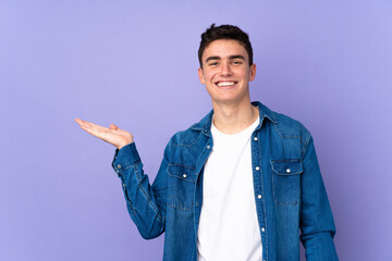 Teenager caucasian  handsome man isolated on purple background holding copyspace imaginary on the...