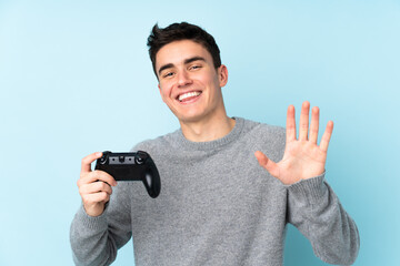 Teenager caucasian man playing with a video game controller isolated on blue background saluting with hand with happy expression