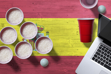 Spain flag concept with plastic beer pong cups and laptop on national wooden table, top view. Beer Pong game.