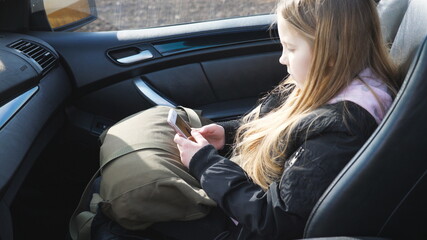 Fototapeta na wymiar Happy little girl with long blonde hair sits in front passenger seat of modern SUV and uses her smart phone. Beautiful female child browses social media on mobile phone, turns to camera and smiles