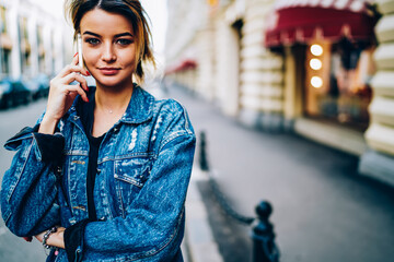 Half length portrait of attractive hipster girl having mobile conversation using application on modern cellular.Cropped image of young woman looking at camera while strolling at urban setting