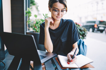 Positive clever hipster girl wear in spectacles and trendy outfit doing homework checking information via modern laptop computer, happy skilled female student writing essay in notebook outdoors