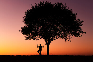 A silhouette of a meditating man next to a tree in front of a sunset