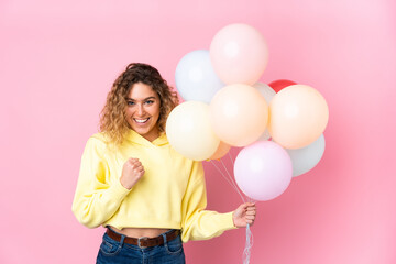 Young blonde woman with curly hair catching many balloons isolated on pink background celebrating a victory