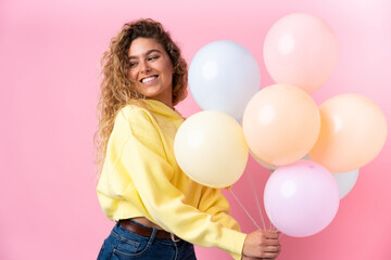 Fototapeta na wymiar Young blonde woman with curly hair catching many balloons isolated on pink background