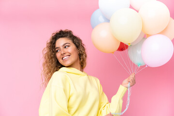 Young blonde woman with curly hair catching many balloons isolated on pink background