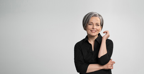 Mature Caucasian woman smiling charmingly with hand near face. Graying elegant woman with short bob hairstyle looks at camera posing on white background. Blank or template with text space at left.