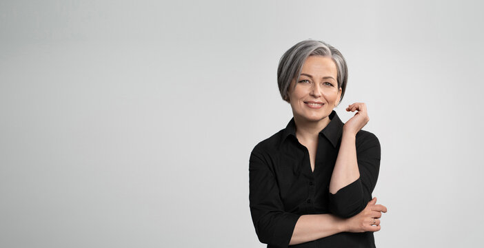 Mature Caucasian woman smiling charmingly with hand near face. Graying elegant woman with short bob hairstyle looks at camera posing on white background. Blank or template with text space at left.