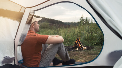 Young man sits near a tent and bonfire against the backdrop of nature. View through the tent. Concept of local travel, tourism and camping. Copy space.