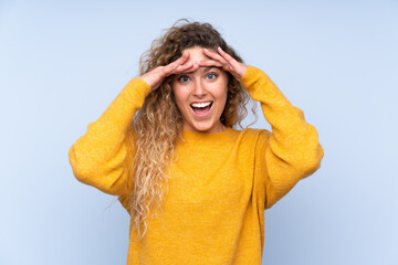 Young blonde woman with curly hair isolated on blue background with surprise expression