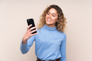 Fototapeta premium Young blonde woman with curly hair wearing a turtleneck sweater isolated on beige background making a selfie