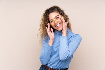 Young blonde woman with curly hair wearing a turtleneck sweater isolated on beige background with glasses and surprised