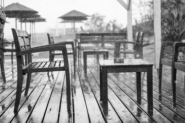 The cafe on the beach bar is empty, there is no one because the weather is bad and it is raining. Wooden chair and table are completely wet. Black and white.