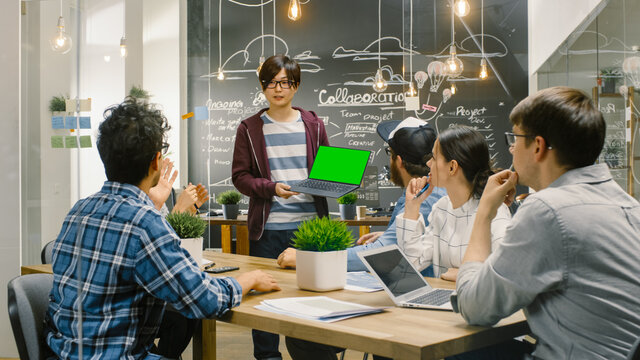 Charismatic Asian Team Leader Shows Laptop With Green Screen Mock-up Template to a Diverse Group of Talented Young Developers. Creative People Have Discussion in Stylish Office Environment.