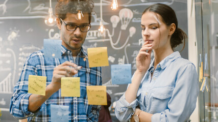 Male and Female Creative Designers Create Outline of the Work Plan with Sticky Papers on Glass Wall and Draw on it with Markers. Stylish Young People in Trendy Environment.