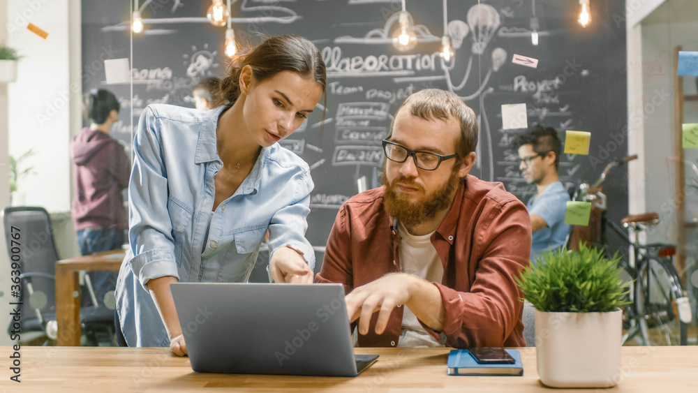Poster female designers talks male developer about ongoing project while he works on a laptop. in the backg