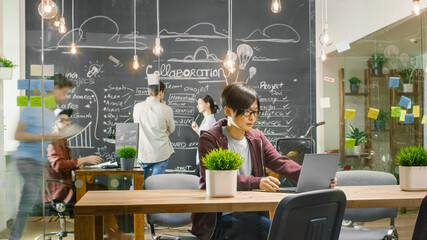 Talented Asian Developer Sits at the Table Works on a Laptop. In the Background Busy Office with...