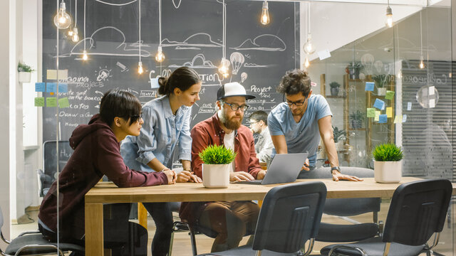 Three Creative People Have Active Discussion About Ongoing Project While Using Laptop. Stylish Young People Work In Trendy Environment.