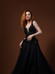 Beautiful elegant young model with bright redhead hairstyle posing in fashion expensive black wedding dress with long skirt on studio bright orange background. Portrait