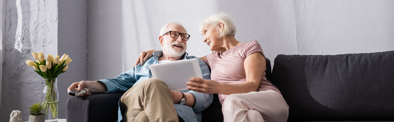 Panoramic shot of smiling elderly woman holding digital tablet near husband with remote controller...