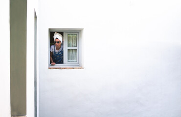 Attractive woman with a towel on her head and sunglasses looking out of a window. White walls.