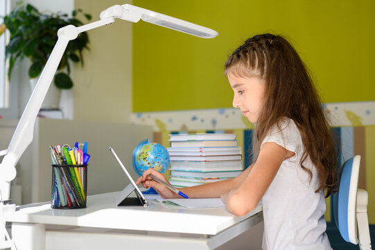A girl sits at a table and uses a tablet. The girl is engaged in remote lessons via the Internet.