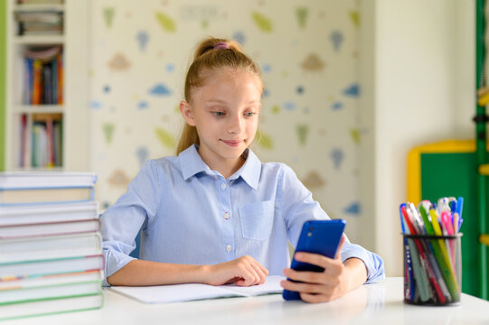A girl sits in a room at a table and holds a smartphone in her hands. Next to the girl is a stack of textbooks.
