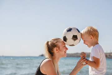 a young blonde mother with a blond son hold a ball between their heads in the summer on the beach....