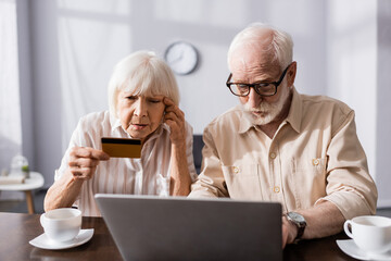 Fototapeta premium Selective focus of senior woman looking at credit card near man using laptop and cups on table