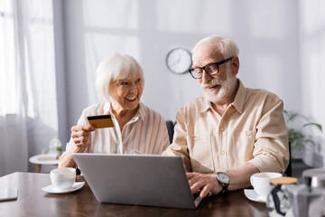 Selective focus of smiling senior couple using laptop and credit card near coffee cups on table