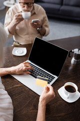 Selective focus of senor woman using laptop and credit card near husband with coffee and smartphone at home
