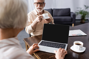 Selective focus of elderly man drinking coffee near wife using laptop at table