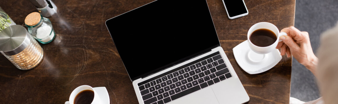 Panoramic Shot Of Elderly Woman Drinking Coffee Near Laptop And Smartphone On Kitchen Table