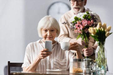 Selective focus of senior woman using smartphone and holding coffee cup near positive man with bouquet at home
