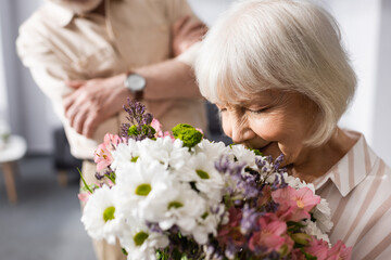 Selective focus of senior woman smelling bouquet near husband at home