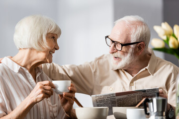 Fototapeta na wymiar Selective focus of senior man holding newspaper and embracing smiling wife with cup of coffee